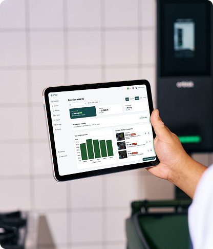 A hand holding an iPad displaying the Orbisk food waste tracking dashboard, with an Orbisk unit visible in the background.