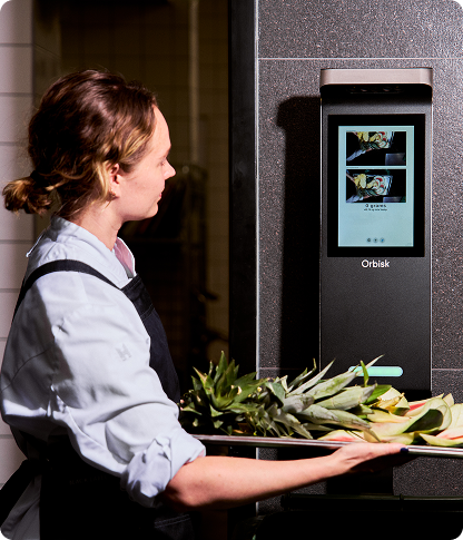 A chef in a grey apron carrying a tray of vegetable trimmings towards an Orbisk food waste monitoring unit mounted on a dark wall.