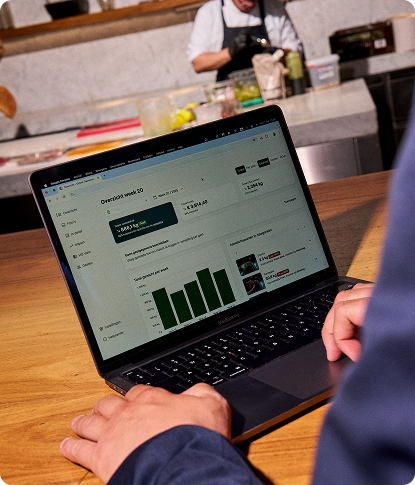 A person reviewing the Orbisk food waste analytics dashboard on a laptop at a restaurant counter, with kitchen staff working in the background.