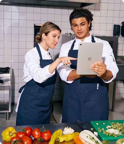 Two chefs in white jackets and navy aprons reviewing data on an iPad in a commercial kitchen, with fresh vegetables on the counter in front of them.