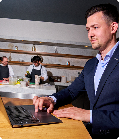 A man in a business suit working on a laptop at a restaurant bar counter, with kitchen staff visible preparing food in the background.