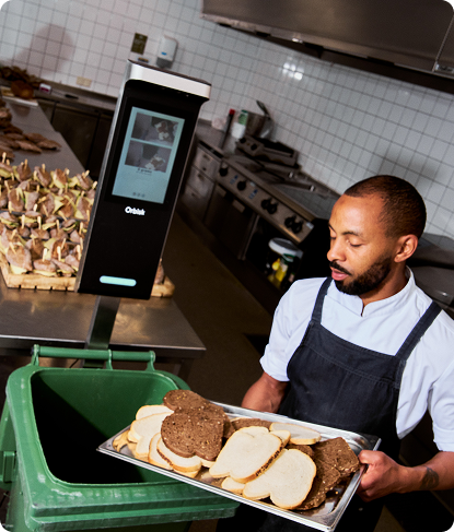 A kitchen worker in a dark apron placing a tray of bread slices near an Orbisk food waste monitoring unit beside a green waste bin.
