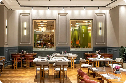 An elegantly appointed hotel restaurant dining room with grey panelled walls, herringbone parquet flooring, and warm track lighting. Tables are set with white linen and glassware, surrounded by wooden chairs with tan and red upholstery. Large interior windows reveal a green-accented bar area beyond.