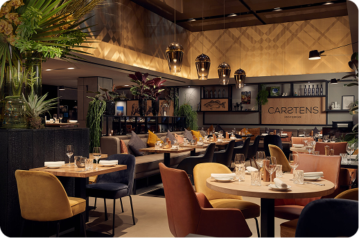 A warmly lit modern restaurant interior featuring round wooden tables with mismatched velvet chairs in mustard, navy, and terracotta. Lush tropical plants hang overhead alongside brushed gold pendant lights. A bar and shelving unit displaying the "Carstens" restaurant name is visible in the background.