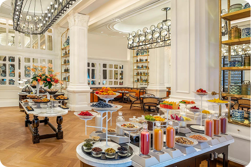 A grand, light-filled breakfast buffet space with high ceilings, white classical columns, and ornate chandeliers. Marble-topped display tables are laden with fresh fruits, juices in glass bottles, and assorted foods. Shelving units line the walls, and the overall aesthetic is elegant and colonial.
