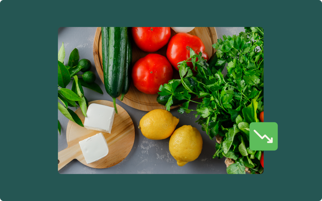 Overhead photo of fresh vegetables and feta cheese on wooden boards, including tomatoes, cucumber, lemons, and herbs, with a downward trend icon overlaid in the corner.