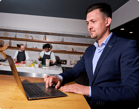orbisk-restaurant-management-laptop (1) Man in pak die een laptop gebruikt in een restaurant, met koks die op de achtergrond eten bereiden
