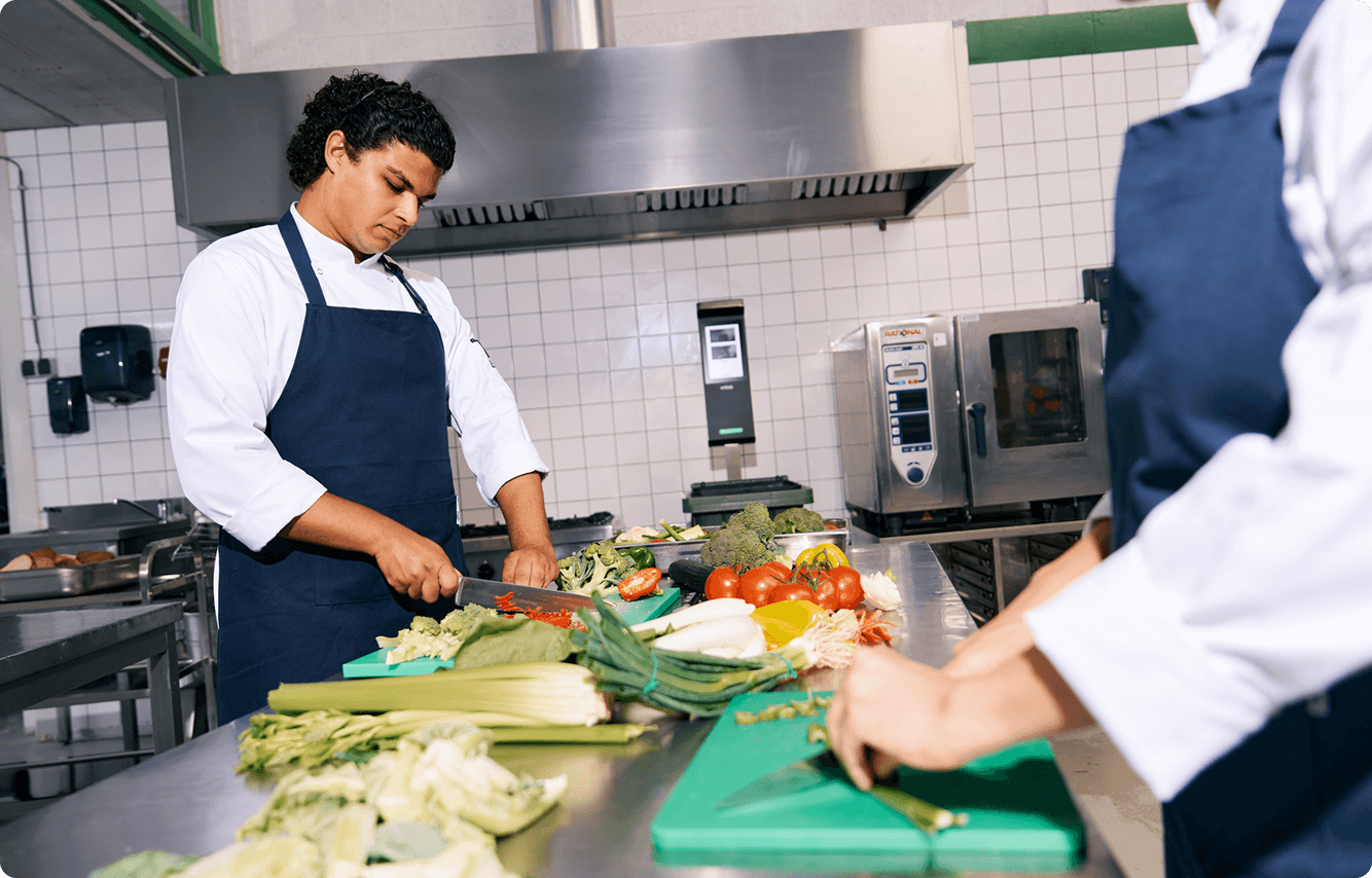 orbisk-chef-vegetable-prep-kitchen Twee koks die groenten bereiden aan de werkbank van een professionele keuken, met een Orbisk-apparaat voor het meten van voedselverspilling aan de muur